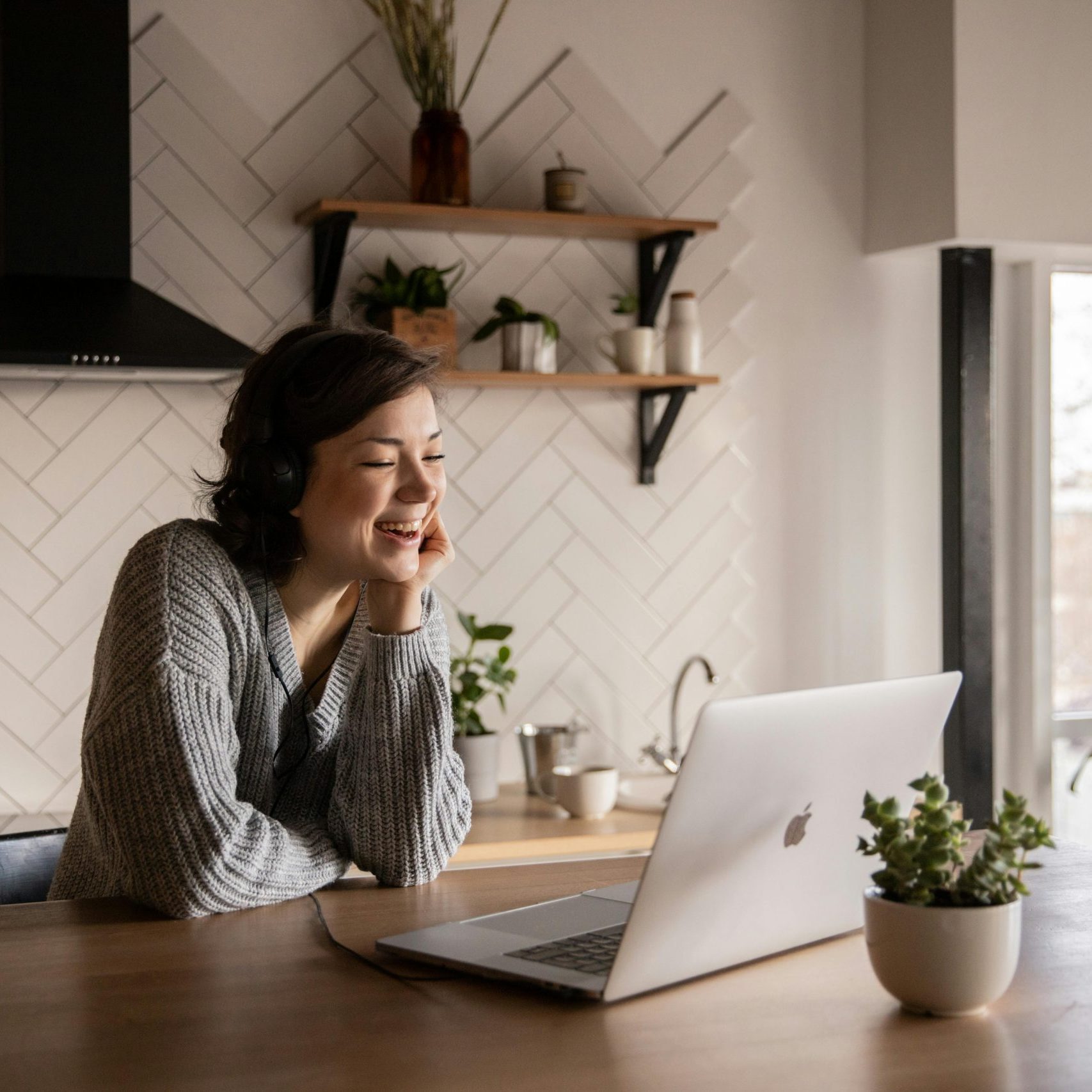 Young cheerful female smiling and talking via laptop while sitting at wooden table in cozy kitchen